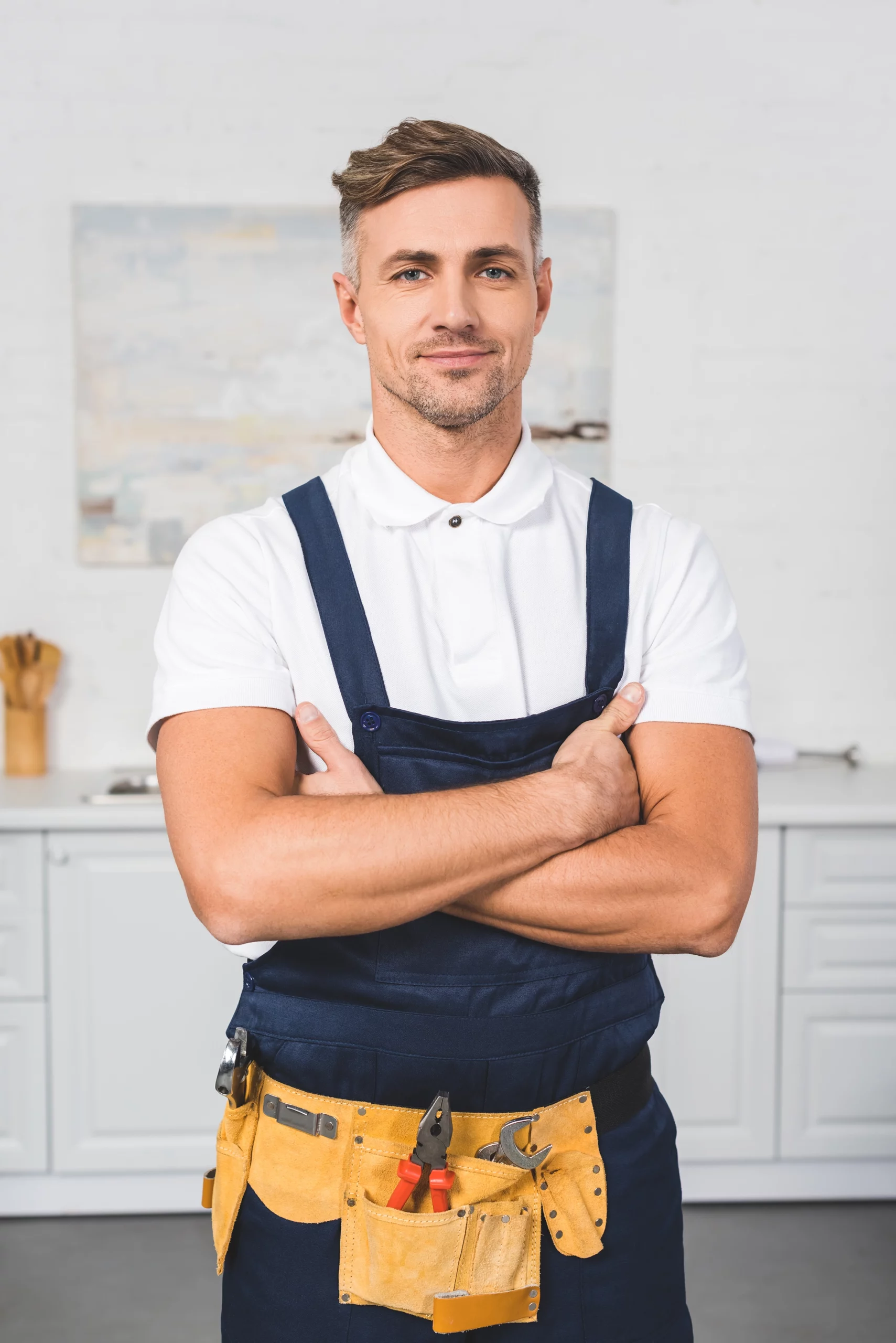 Un professionnel de la réparation, ceinture à outils autour de la taille, se tient debout dans la cuisine, les bras croisés, fixant la caméra.