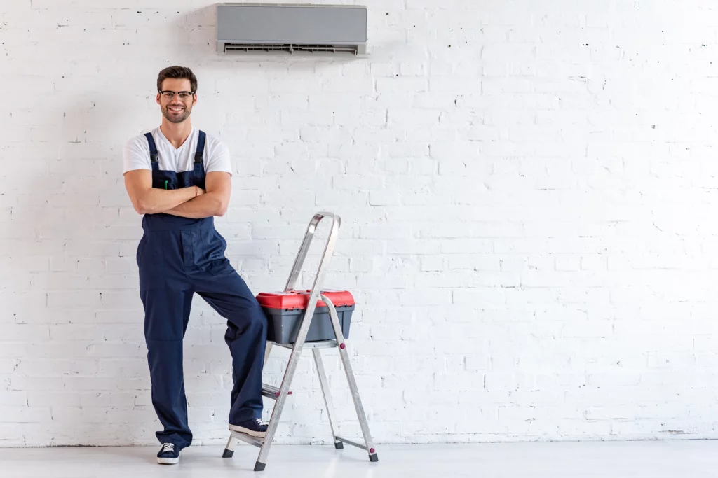 Réparateur souriant debout sous le climatiseur près d'un escabeau et d'une boîte à outils, regardant la caméra.